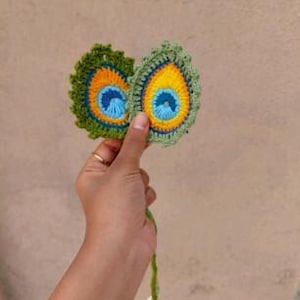 Peut inclure: Deux appliqués faits à la main en crochet en forme de plumes de paon. Les plumes sont vertes, jaunes, bleues et oranges. Une gland vert avec des perles blanches est attaché aux plumes. Les appliqués sont tenus dans une main.