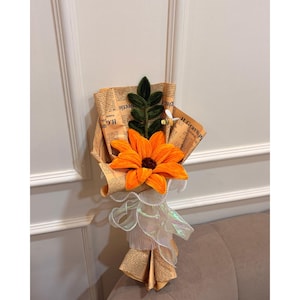 Peut inclure: Un bouquet fait à la main avec un grand tournesol orange comme point central. La fleur est entourée de feuilles vertes et d'une petite abeille. Le bouquet est enveloppé dans du papier journal vintage et noué avec un ruban blanc.