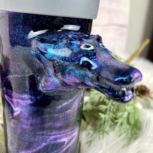 May include: A tumbler with a galaxy-themed design in shades of purple, blue, and black. A sculpted dragon head is attached to the side. The tumbler has a gray lid and is set against a soft, white background.