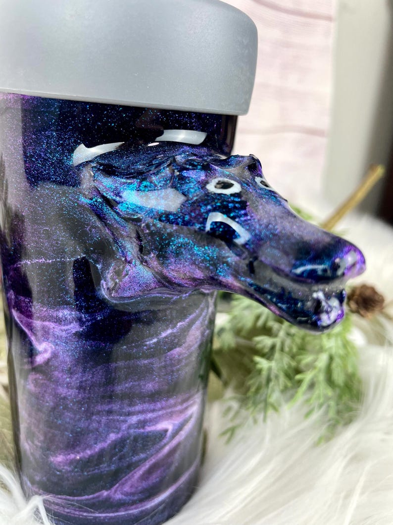 May include: A tumbler with a galaxy-themed design in shades of purple, blue, and black. A sculpted dragon head is attached to the side. The tumbler has a gray lid and is set against a soft, white background.
