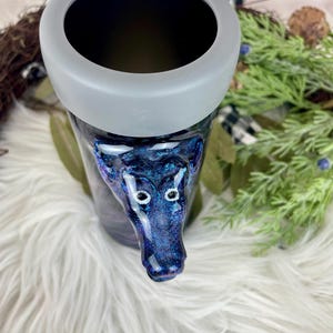 May include: A tumbler with a dark blue and purple glitter design, featuring a wolf head. The tumbler has a gray rim and a black interior. The wolf head is detailed with white eyes. The tumbler sits on a white, fluffy surface.