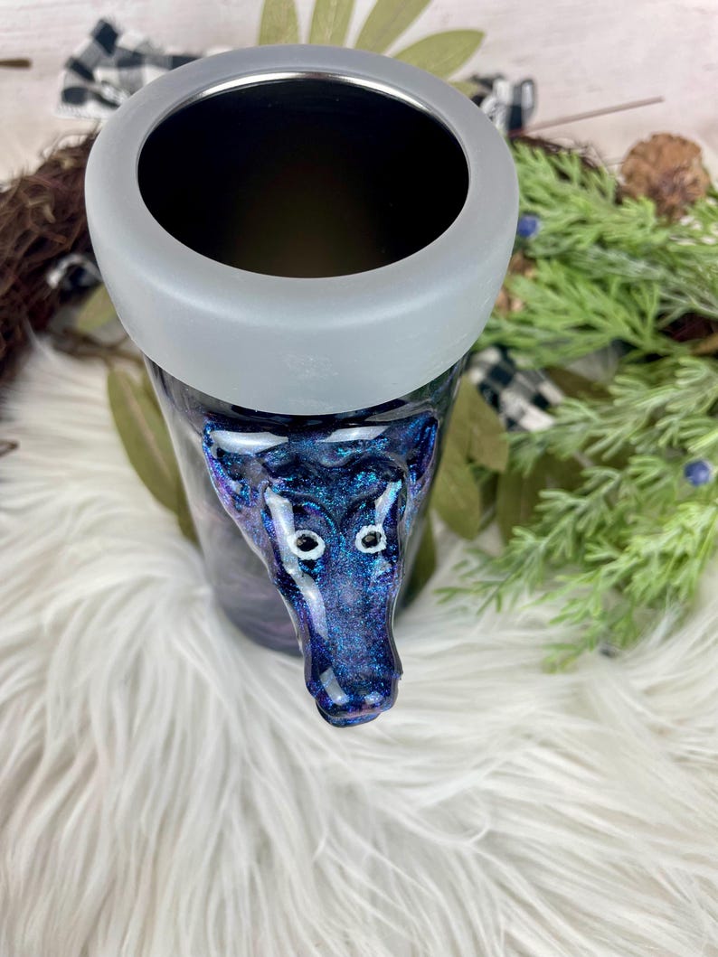 May include: A tumbler with a dark blue and purple glitter design, featuring a wolf head. The tumbler has a gray rim and a black interior. The wolf head is detailed with white eyes. The tumbler sits on a white, fluffy surface.