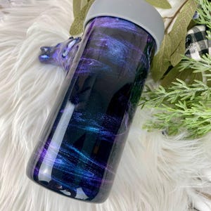 May include: A stainless steel tumbler with a galaxy-themed design. The tumbler features a dark background with swirling shades of purple, blue, and teal. It has a gray lid and a smooth, glossy finish. The tumbler is designed to keep beverages hot or cold.