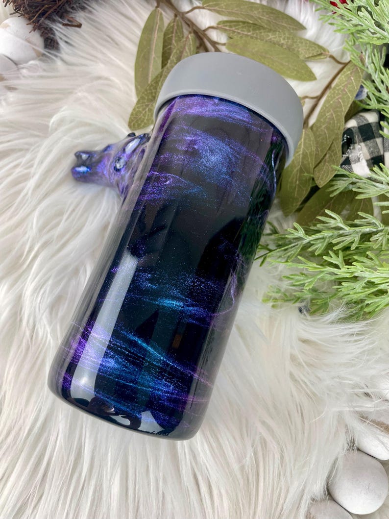 May include: A stainless steel tumbler with a galaxy-themed design. The tumbler features a dark background with swirling shades of purple, blue, and teal. It has a gray lid and a smooth, glossy finish. The tumbler is designed to keep beverages hot or cold.