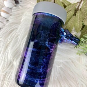 May include: A cylindrical tumbler with a galaxy-themed design in shades of blue, purple, and silver. The tumbler has a gray lid and a decorative handle. The design resembles a nebula or starry night.