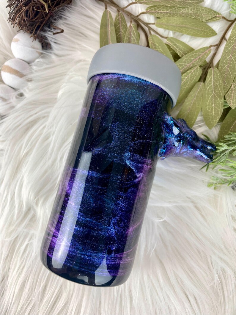 May include: A cylindrical tumbler with a galaxy-themed design in shades of blue, purple, and silver. The tumbler has a gray lid and a decorative handle. The design resembles a nebula or starry night.