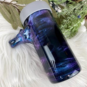 May include: A tumbler with a horse head handle, featuring a swirling galaxy design in shades of purple, blue, and black. The tumbler has a gray rim and is set against a white, fluffy background. The horse head handle adds a unique touch.