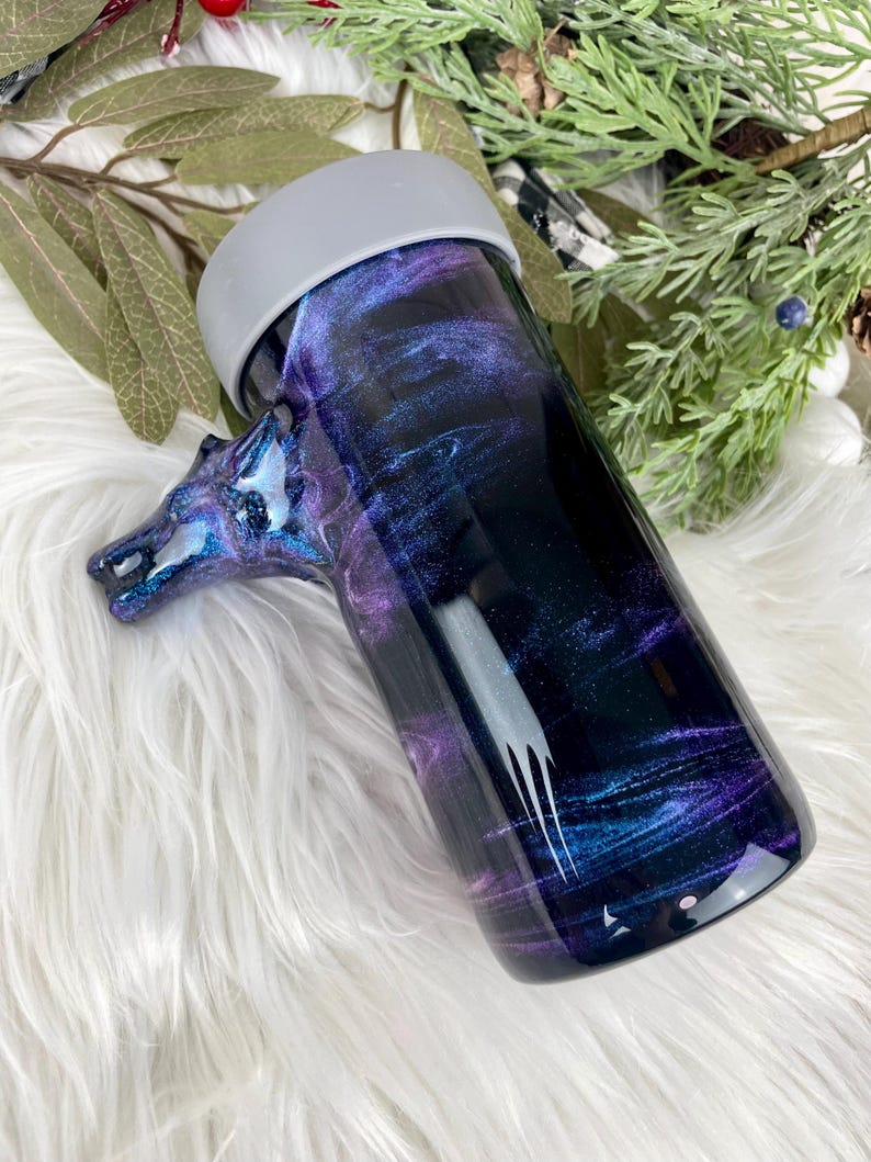 May include: A tumbler with a horse head handle, featuring a swirling galaxy design in shades of purple, blue, and black. The tumbler has a gray rim and is set against a white, fluffy background. The horse head handle adds a unique touch.