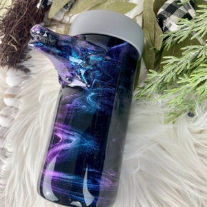May include: A tumbler with a wolf head design. The tumbler features a swirling galaxy design in shades of purple, blue, and black. The lid is gray. The wolf head is a dark color and is part of the tumbler's design.