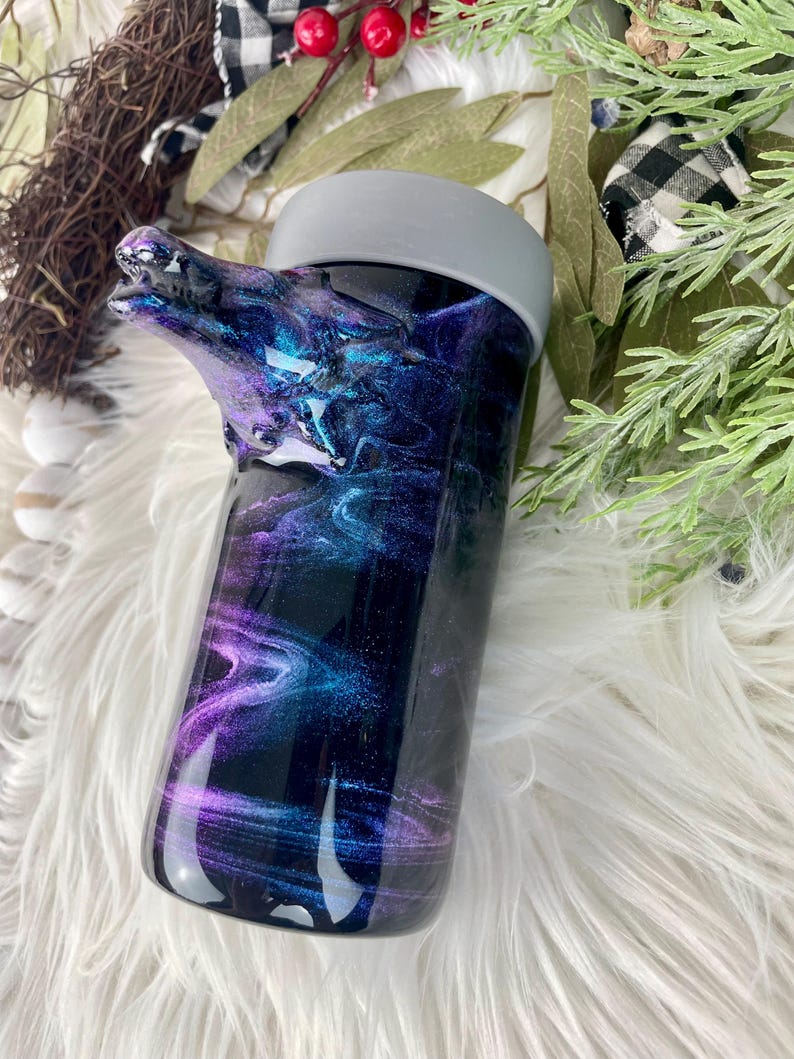 May include: A tumbler with a wolf head design. The tumbler features a swirling galaxy design in shades of purple, blue, and black. The lid is gray. The wolf head is a dark color and is part of the tumbler's design.