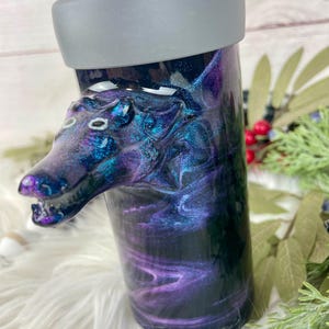 May include: A tumbler with a wolf head design. The tumbler has a swirling galaxy pattern in shades of purple, blue, and black. The wolf head is a detailed sculpture with white accents. The lid is gray.