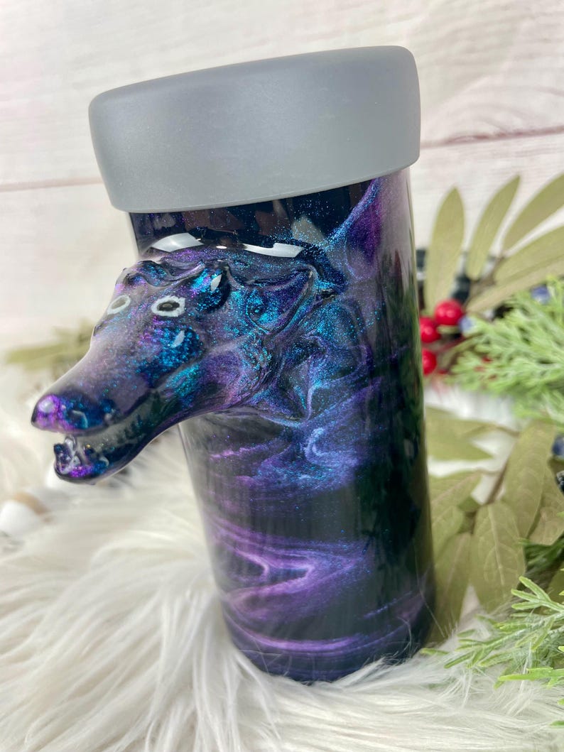May include: A tumbler with a wolf head design. The tumbler has a swirling galaxy pattern in shades of purple, blue, and black. The wolf head is a detailed sculpture with white accents. The lid is gray.