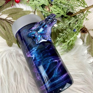 May include: A stainless steel tumbler with a galaxy-themed design in shades of blue, purple, and black. The tumbler features a sculpted wolf head on the side and a gray lid. The design has a swirling, cosmic effect.