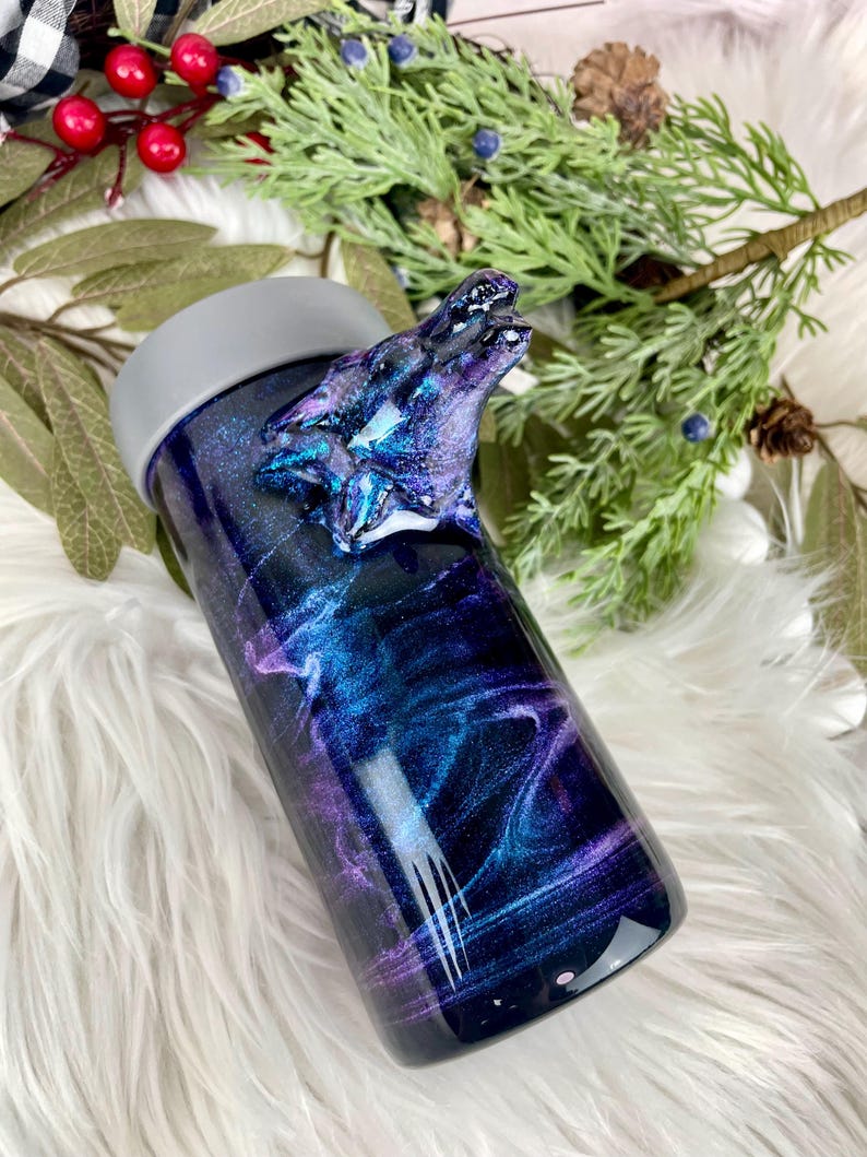 May include: A stainless steel tumbler with a galaxy-themed design in shades of blue, purple, and black. The tumbler features a sculpted wolf head on the side and a gray lid. The design has a swirling, cosmic effect.
