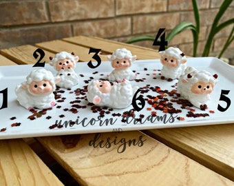 May include: Six white fluffy sheep figurines with pink accents, numbered 1 through 6, on a white tray with brown glitter stars. The tray has the text "unicorn dreams and designs" on it.