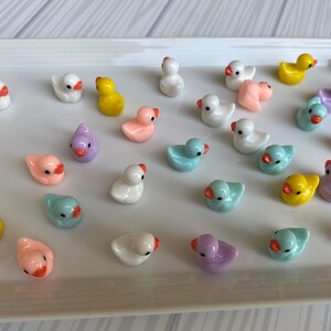 May include: A collection of miniature ceramic duck figurines in various pastel colors, including yellow, pink, blue, purple, and white. Each duck has a small orange beak and black eyes. The ducks are arranged on a white rectangular tray.