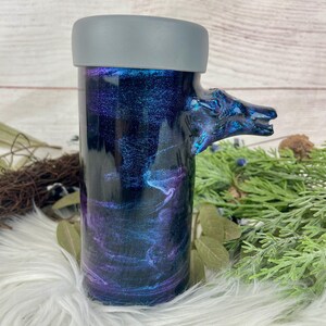May include: A cylindrical tumbler with a gray lid and a handle shaped like a wolf's head. The tumbler features a swirling design in shades of blue, purple, and black. The handle is a darker blue with iridescent accents.