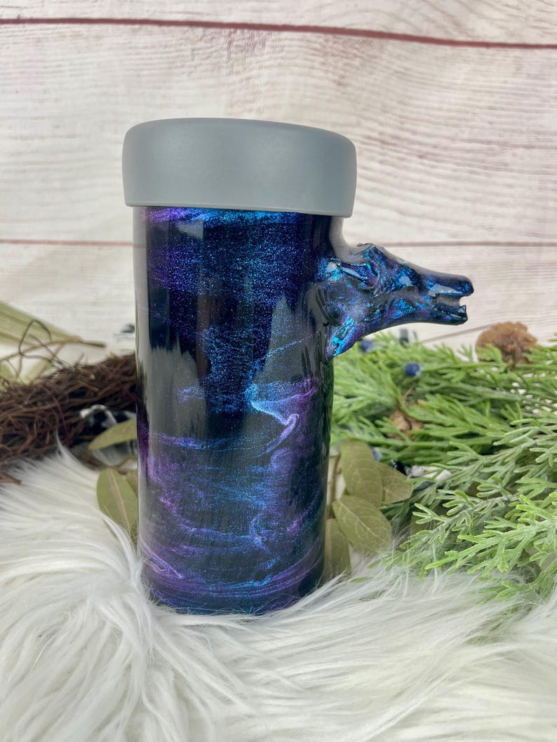 May include: A cylindrical tumbler with a gray lid and a handle shaped like a wolf's head. The tumbler features a swirling design in shades of blue, purple, and black. The handle is a darker blue with iridescent accents.