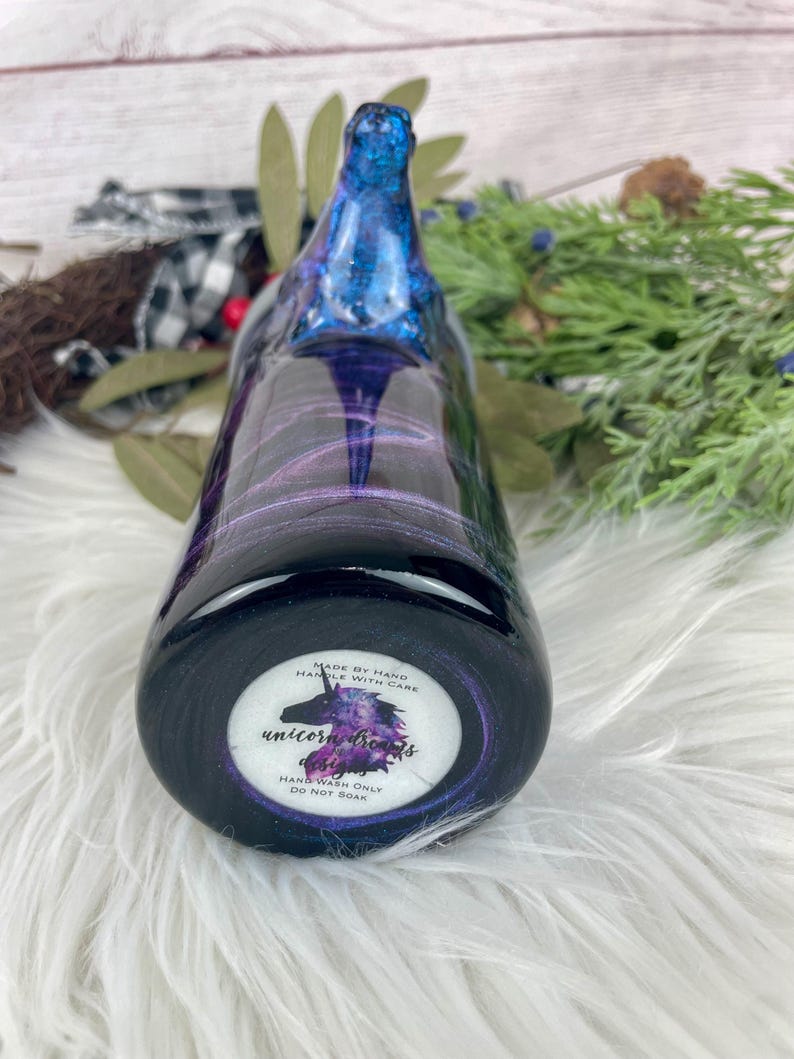 May include: A handcrafted, dark-colored bottle with a swirling blue and purple design. The bottle's base features a circular label with a unicorn illustration and the text "Unicorn Dreams Designs". The label also includes the text "Hand Wash Only, Do Not Soak".