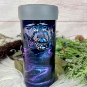 May include: A tumbler with a gray top and a swirling blue and purple design. The tumbler features a sculpted bear head. The design resembles a galaxy. The tumbler is cylindrical and appears to be made of resin.