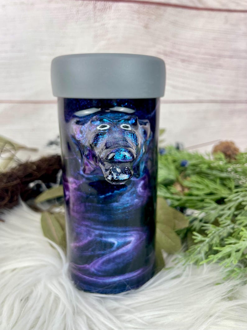 May include: A tumbler with a gray top and a swirling blue and purple design. The tumbler features a sculpted bear head. The design resembles a galaxy. The tumbler is cylindrical and appears to be made of resin.