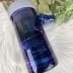 May include: A tumbler with a swirling galaxy design in shades of purple, blue, and black. The tumbler has a gray lid and a handle shaped like a wolf's head. The design is a mix of glitter and resin.