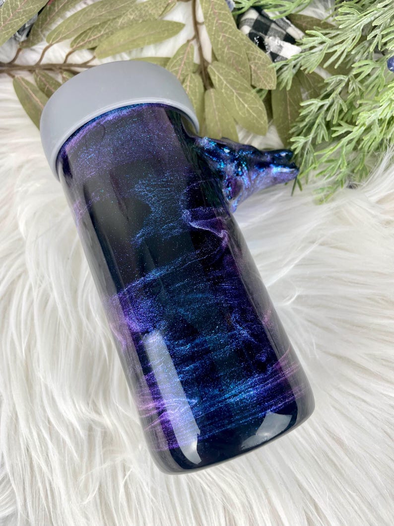 May include: A tumbler with a swirling galaxy design in shades of purple, blue, and black. The tumbler has a gray lid and a handle shaped like a wolf's head. The design is a mix of glitter and resin.