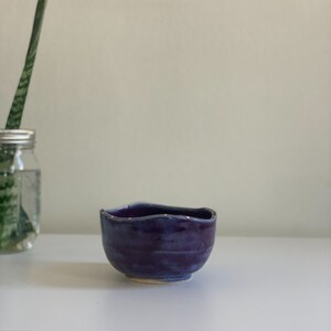 Puede incluir: Un pequeño cuenco de cerámica morado con un borde ondulado y detalles dorados. El cuenco está sobre una superficie blanca.