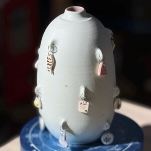 Puede incluir: Un jarrón de cerámica blanco roto hecho a mano, con forma bulbosa y una pequeña abertura. El jarrón está adornado con pequeños amuletos colgantes, incluyendo un pez, un corazón y una carta de juego. El jarrón está sobre una superficie azul.