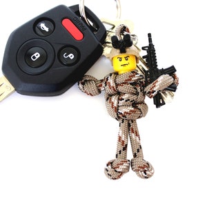 K&ouml;nnte beinhalten: Ein Schl&uuml;sselanh&auml;nger aus Tarn-Paracord mit einer Lego-Minifigur, die ein schwarzes Gewehr h&auml;lt. Die Minifigur hat ein gelbes Gesicht mit einem schwarzen Helm und einem schwarzen und braunen Tarnmuster.
