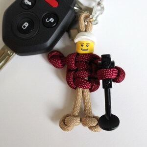 May include: A maroon paracord keychain with a Lego minifigure head and a black Lego piece attached. The minifigure has a yellow face with a smile and a white helmet.