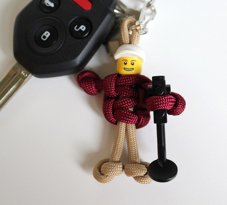 May include: A maroon paracord keychain with a Lego minifigure head and a black Lego piece attached. The minifigure has a yellow face with a smile and a white helmet.