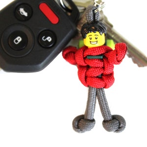 May include: A red and gray paracord keychain with a Lego minifigure head. The minifigure has a yellow face with a smile and black hair.