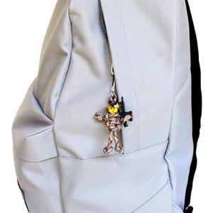 K&ouml;nnte beinhalten: Ein grauer Rucksack mit einer Rei&szlig;verschlusstasche auf der Vorderseite. An dem Rei&szlig;verschlussanh&auml;nger ist eine kleine Tarnfigur aus Paracord mit einem gelben Kopf und einer schwarzen Pistole befestigt.