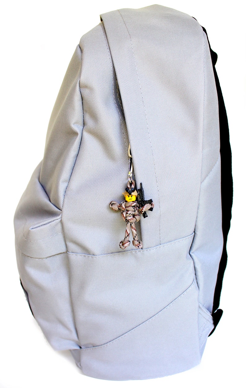 K&ouml;nnte beinhalten: Ein grauer Rucksack mit einer Rei&szlig;verschlusstasche auf der Vorderseite. An dem Rei&szlig;verschlussanh&auml;nger ist eine kleine Tarnfigur aus Paracord mit einem gelben Kopf und einer schwarzen Pistole befestigt.
