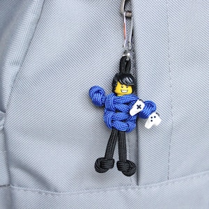 May include: A blue and black paracord keychain with a small figure holding a video game controller. The figure has a yellow face with a black smile and black hair.