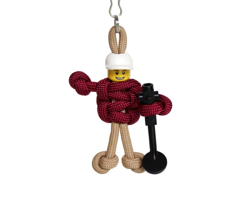May include: A red and tan paracord figure of a Lego minifigure wearing a white hard hat and holding a black tool.