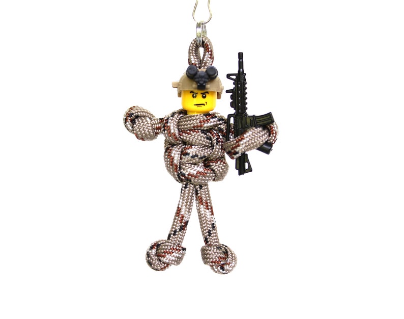 K&ouml;nnte beinhalten: Ein Paracord-Schl&uuml;sselanh&auml;nger mit einer Figur eines Soldaten in Tarnmuster, der ein schwarzes Gewehr h&auml;lt. Die Figur hat einen gelben Lego-Kopf mit einem schwarzen Helm und einer schwarzen Schutzbrille.
