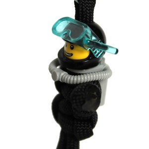 May include: A black and yellow Lego minifigure wearing a black scuba diving helmet and a blue diving mask. The minifigure is attached to a black paracord.