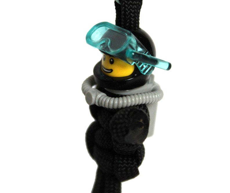 May include: A black and yellow Lego minifigure wearing a black scuba diving helmet and a blue diving mask. The minifigure is attached to a black paracord.