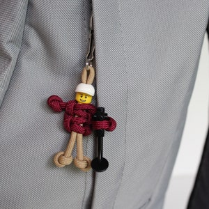 May include: A small, yellow Lego figure with a white helmet is attached to a zipper pull on a gray backpack. The figure is holding a black tool and is made of red, brown, and tan paracord.