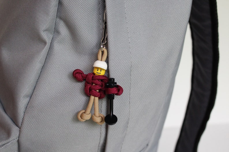 May include: A small, yellow Lego figure with a white helmet is attached to a zipper pull on a gray backpack. The figure is holding a black tool and is made of red, brown, and tan paracord.