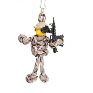 K&ouml;nnte beinhalten: Ein Schl&uuml;sselanh&auml;nger aus Paracord, der eine Soldatenfigur mit Helm und einem schwarzen Gewehr darstellt. Die Figur ist aus braunem und beigefarbenem Paracord gefertigt.