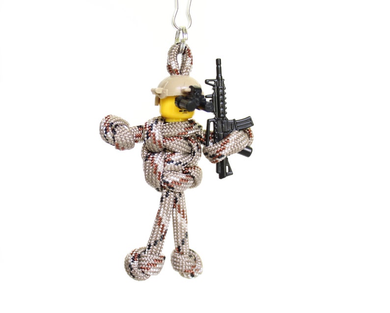 K&ouml;nnte beinhalten: Ein Schl&uuml;sselanh&auml;nger aus Paracord, der eine Soldatenfigur mit Helm und einem schwarzen Gewehr darstellt. Die Figur ist aus braunem und beigefarbenem Paracord gefertigt.