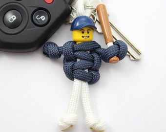May include: A blue and white paracord keychain with a Lego figure holding a brown baseball bat. The figure is wearing a blue baseball cap.