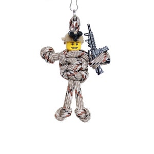 K&ouml;nnte beinhalten: Eine kleine, braune und beigefarbene Paracord-Figur eines Soldaten, der eine schwarze Spielzeugpistole h&auml;lt. Die Figur tr&auml;gt einen gelben Helm mit einem schwarzen Gesicht.