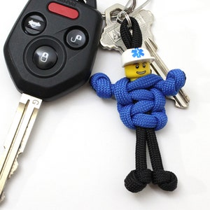 May include: A blue and black paracord keychain with a small white and yellow figure wearing a blue uniform and a white hat with a red cross. The figure is attached to a silver key chain with a black fob.