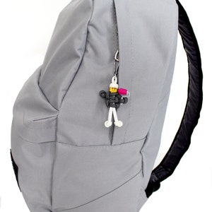 May include: A gray backpack with a black zipper and a small white and yellow figure keychain attached to the zipper pull.