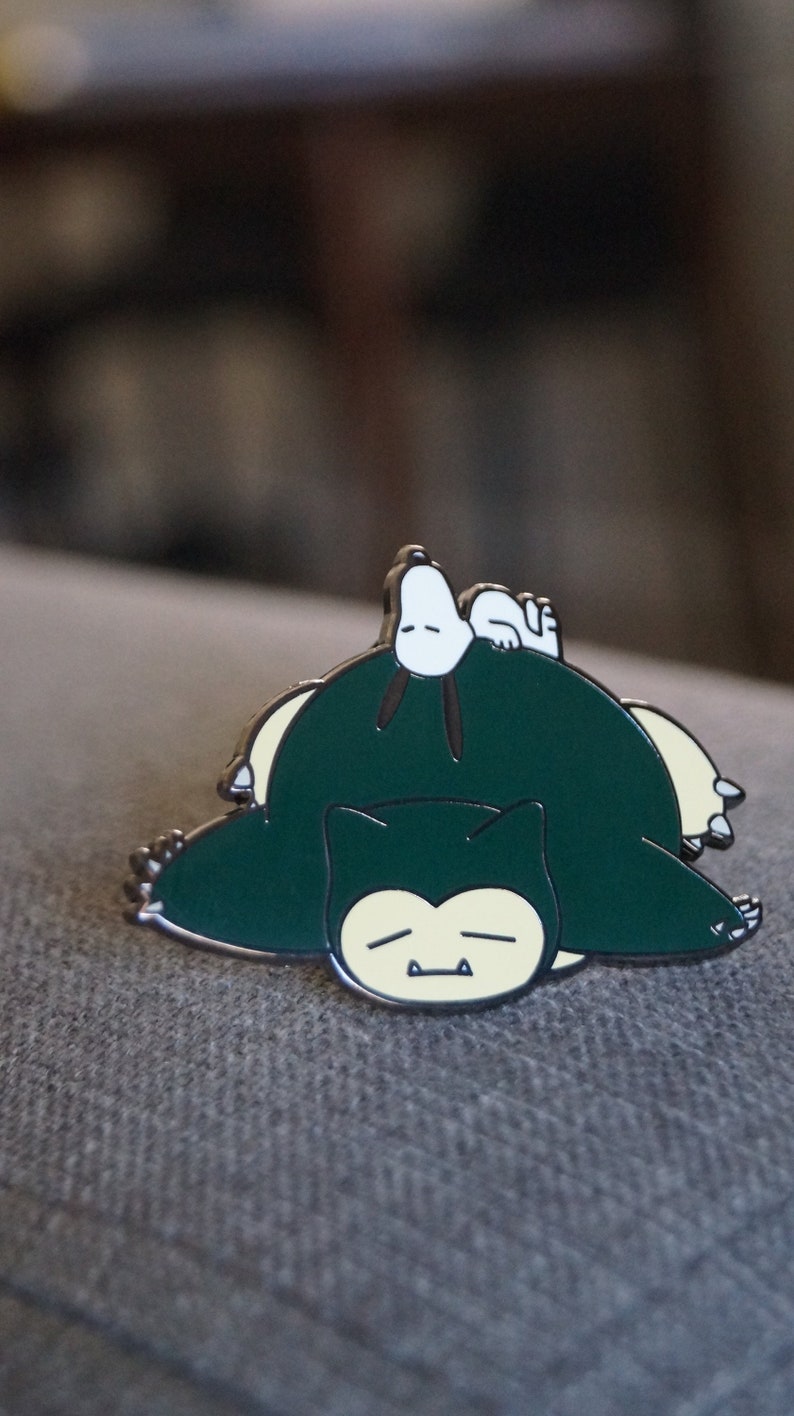 Op de afbeelding: Een groene en gele emaille pin met een cartoonfiguur uit de Pok&eacute;mon-franchise, Snorlax, die slaapt met een witte cartoonhond erop.