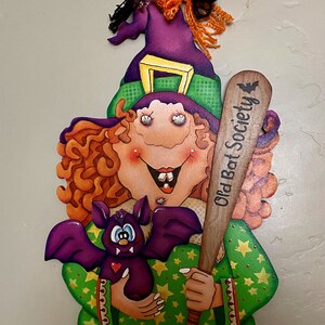 May include: A green and purple witch with orange hair holds a purple bat and a wooden bat with the text "Old Bat Society".
